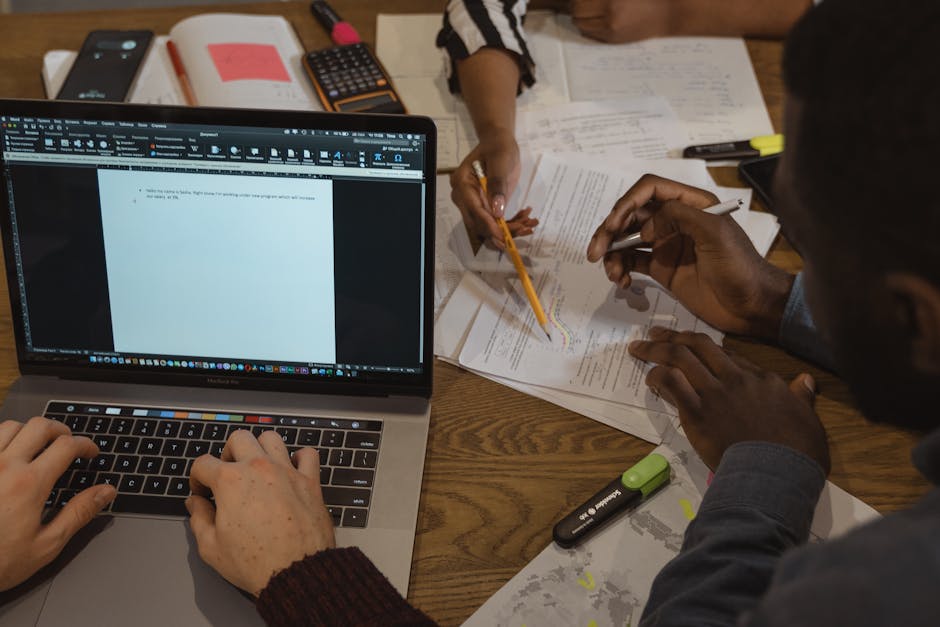 Diverse team collaborating in an office, working on laptops and documents around a table.